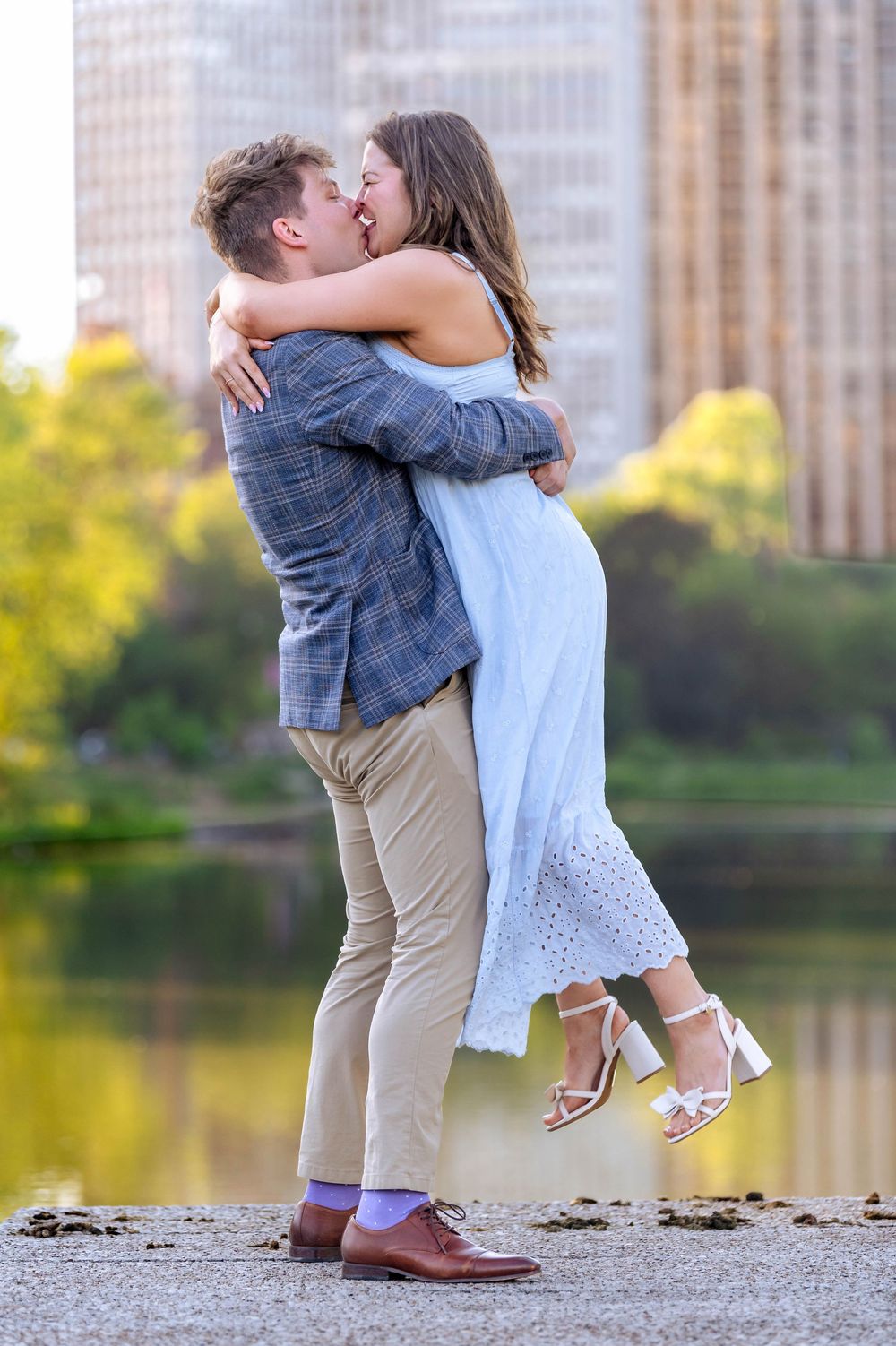 Couple embracing and kissing by a lake in Central Park, man lifting woman in white dress and heels.