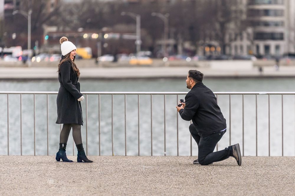 Man proposing on one knee to woman by waterfront railing in winter, holding ring box in romantic outdoor setting.