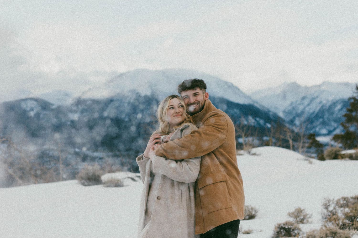 Engagement couple in warm winter coats while standing on a snowy mountain peak with misty Breckenridge, Colorado mountains in background.