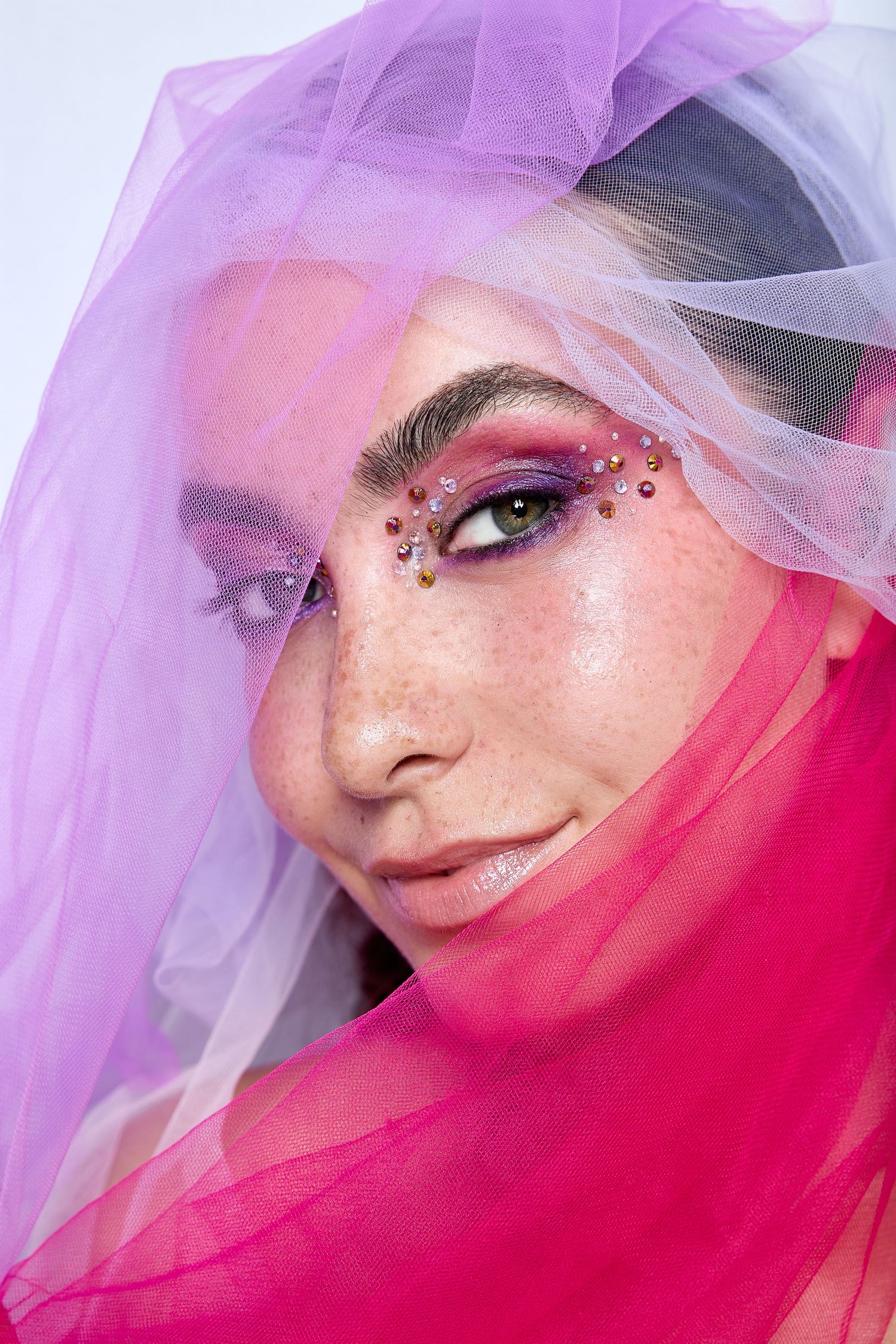 Close-up of woman with bold gem-studded eye makeup, framed by pink and purple tulle for a vibrant, artistic beauty shot.