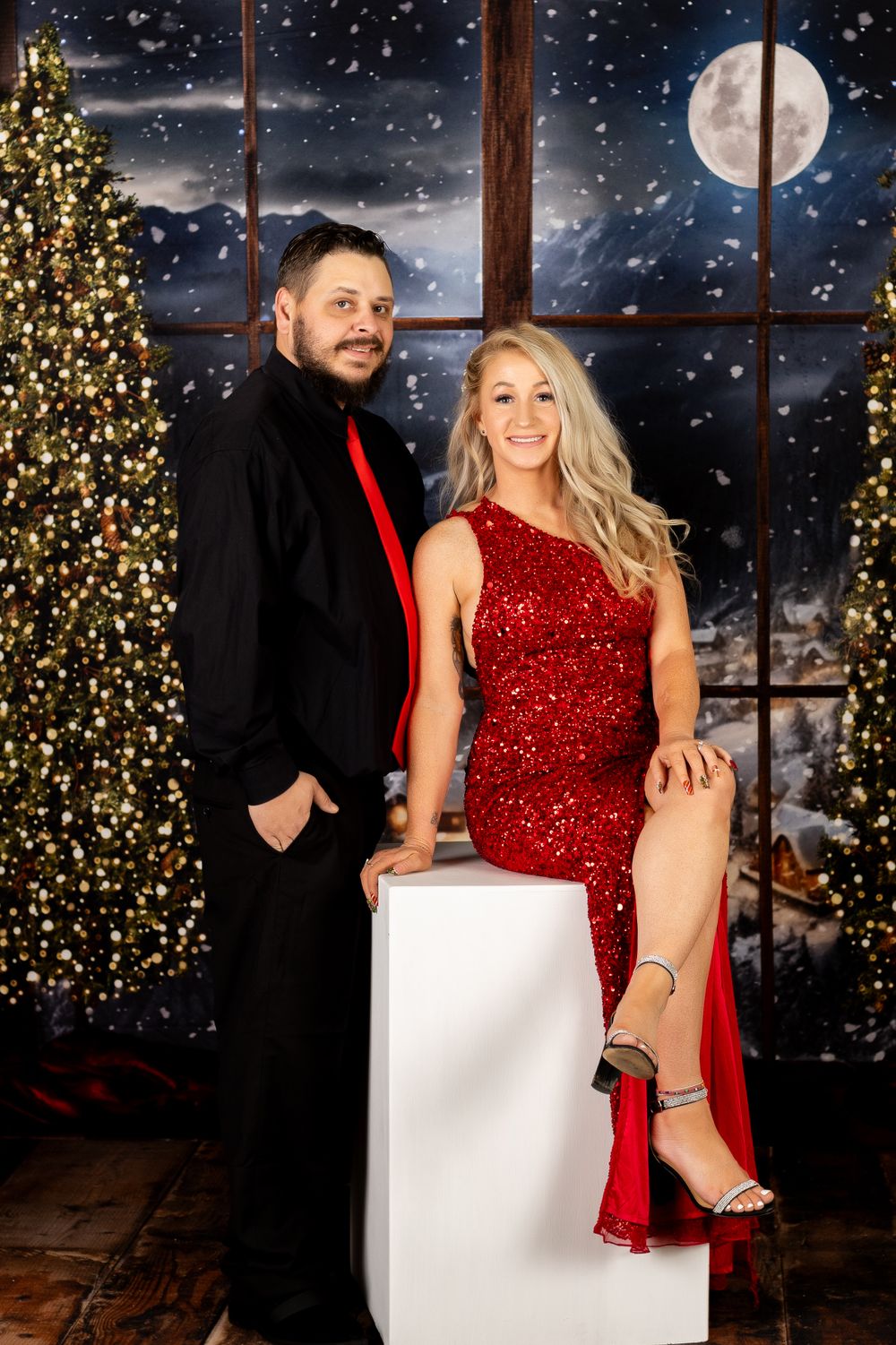 Couple in festive attire posing before Christmas tree backdrop with snowy night scene behind them.