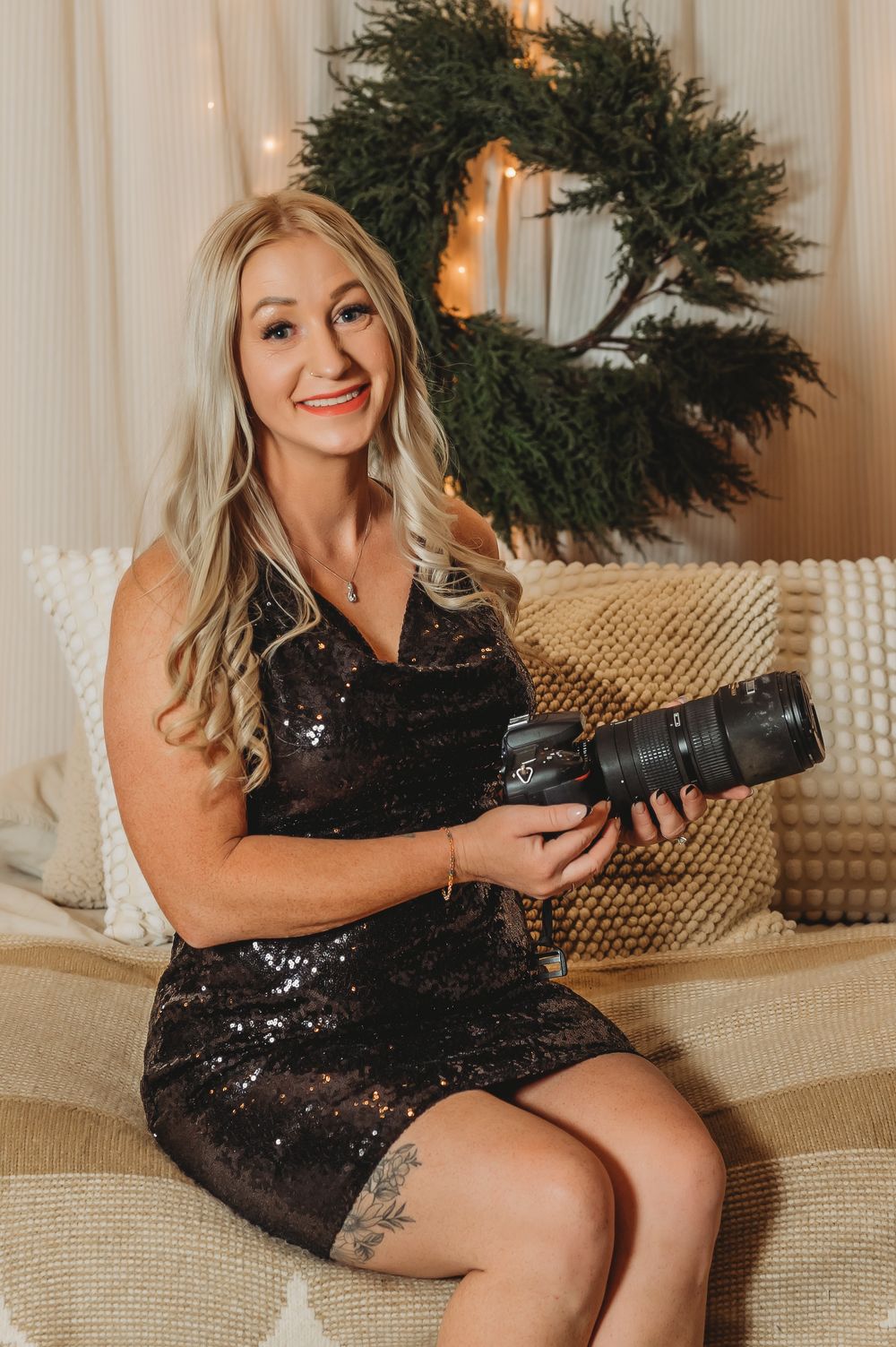 A blonde female photographer in a black sequin dress sits on a couch holding a camera near a holiday wreath.
