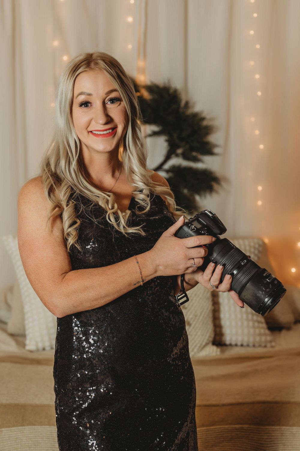 A smiling blonde female photographer in a black sequin dress holds a DSLR camera with fairy lights behind her.
