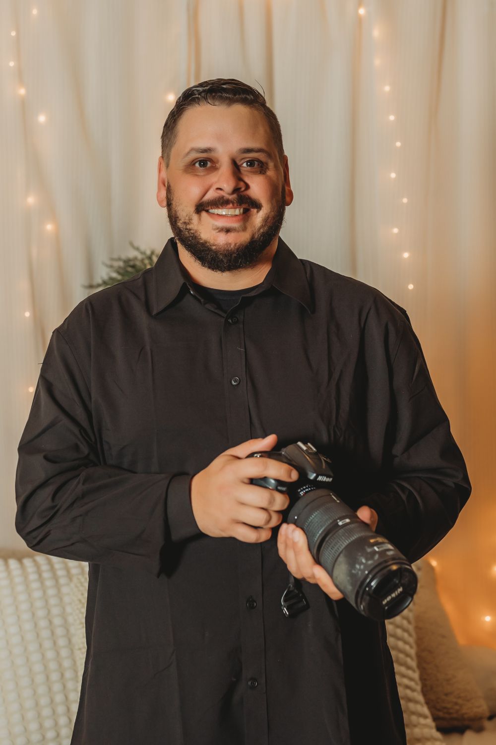 A smiling male photographer in a black shirt holds a DSLR camera against a warm bokeh light backdrop.