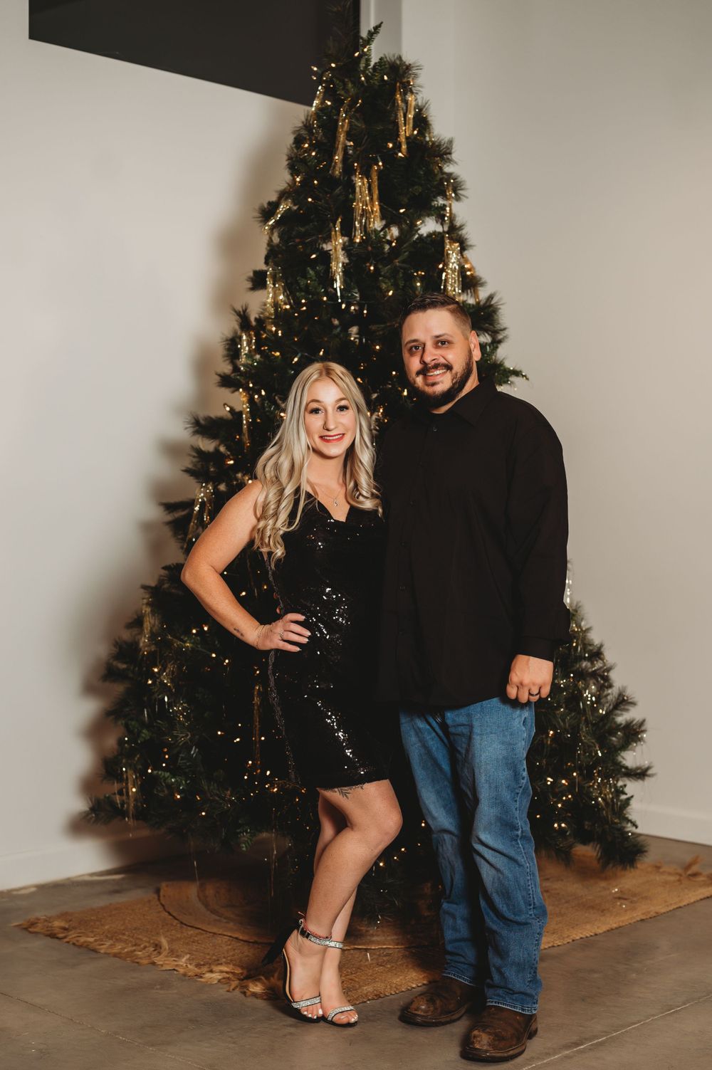 A couple poses together in front of a decorated Christmas tree, dressed in elegant black holiday attire.