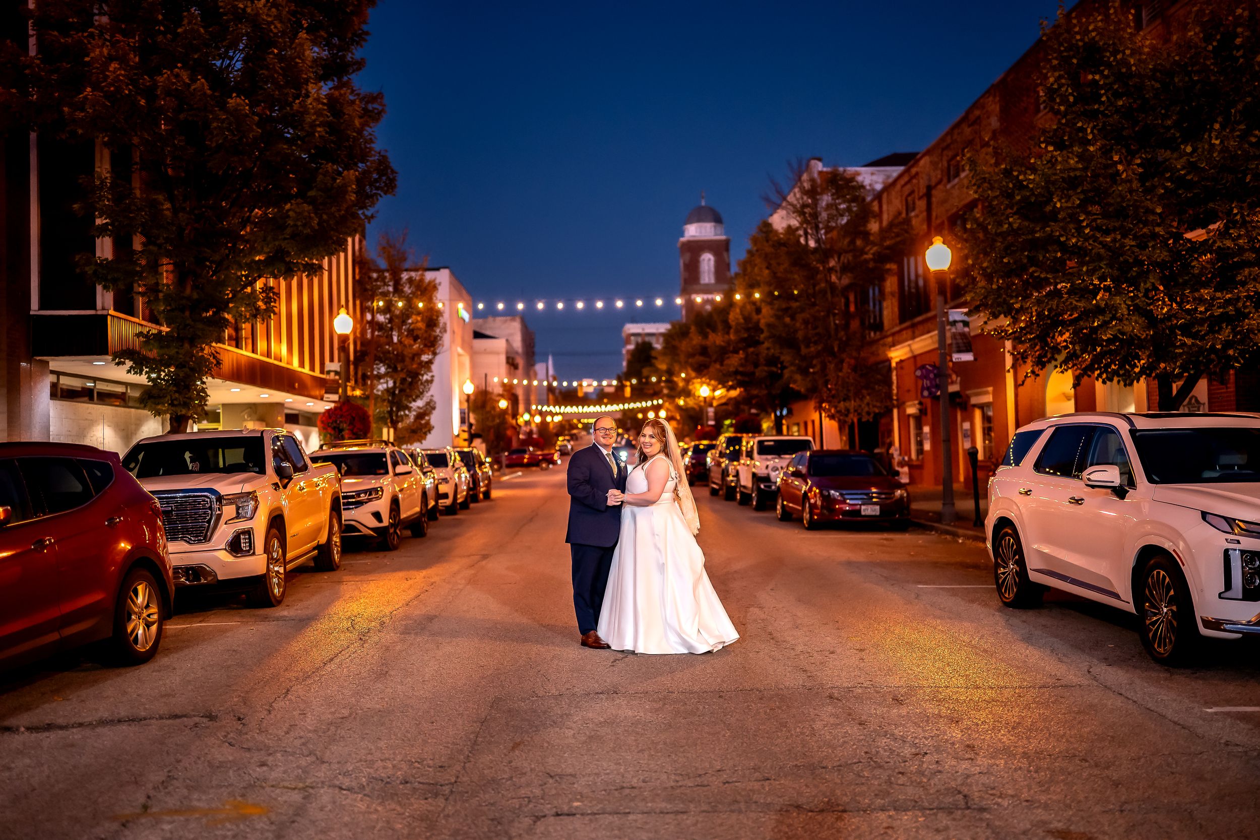 A couple stands in the middle of a downtown street at night with string lights overhead and cars parked along both sides.