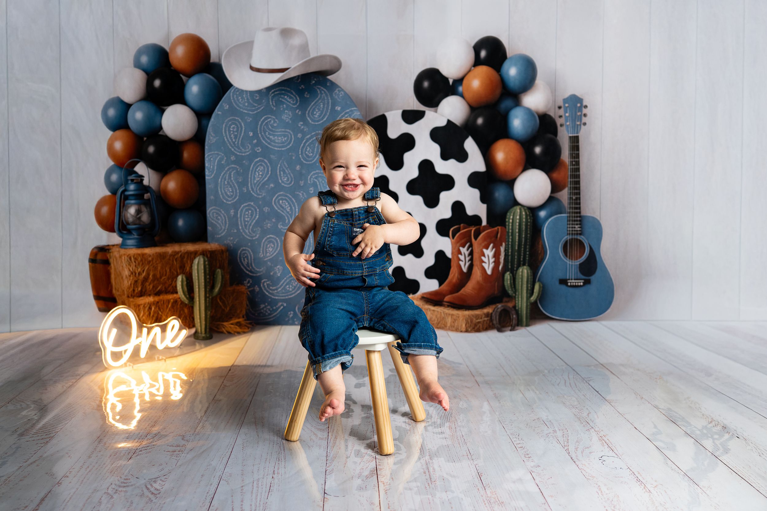 Smiling baby boy in denim overalls sits on stool at western-themed first birthday party with balloons and guitar.