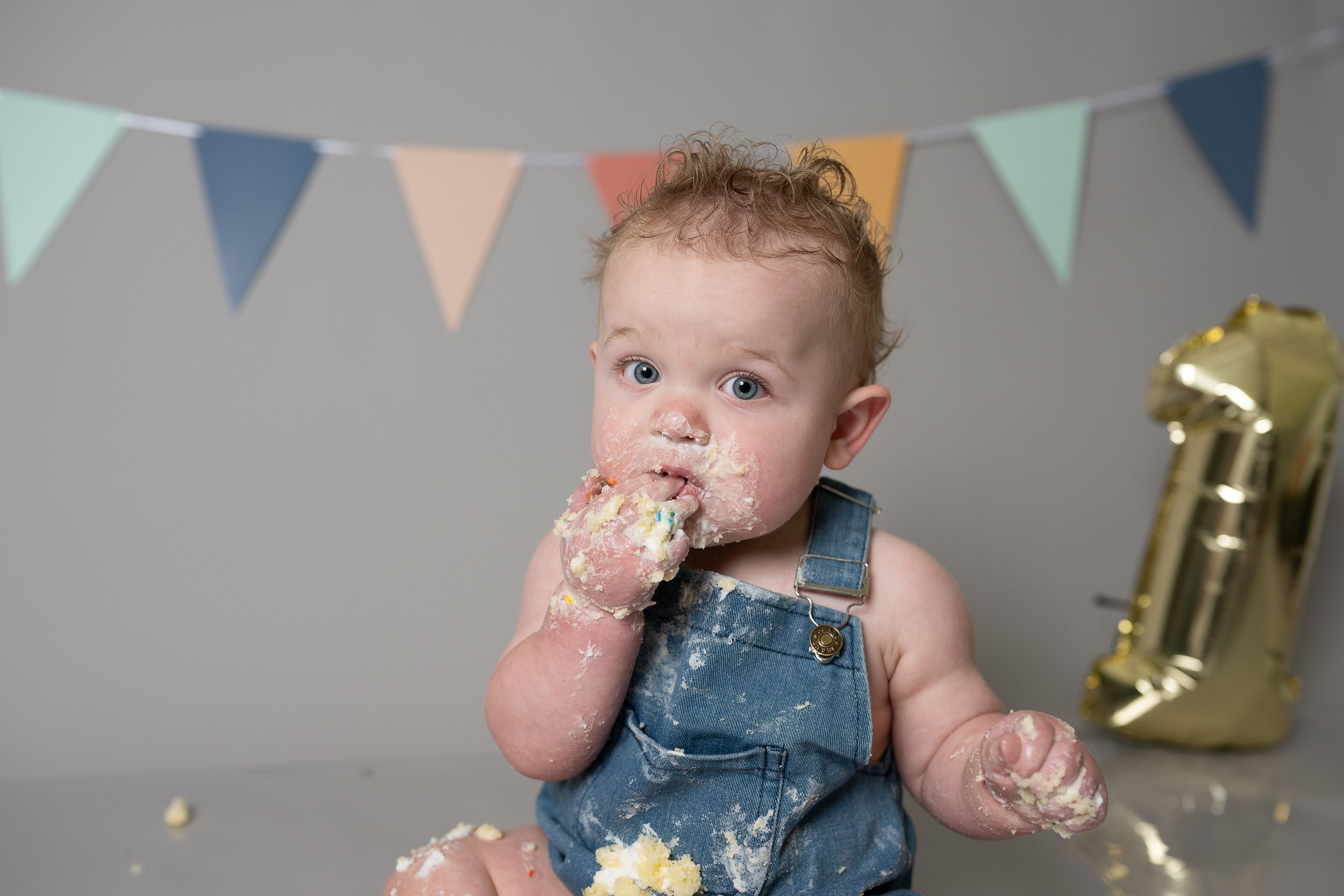 Baby in denim overalls eating cake during first birthday smash cake photoshoot with colorful bunting.