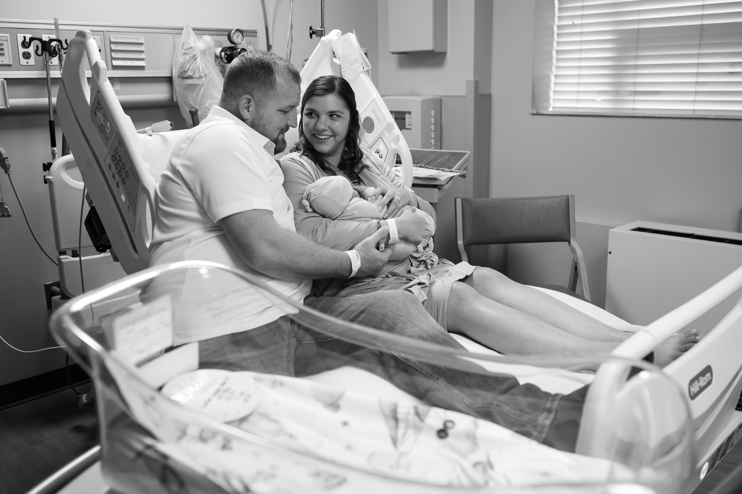 Black and white photo of new parents holding their newborn baby in a hospital room after delivery.