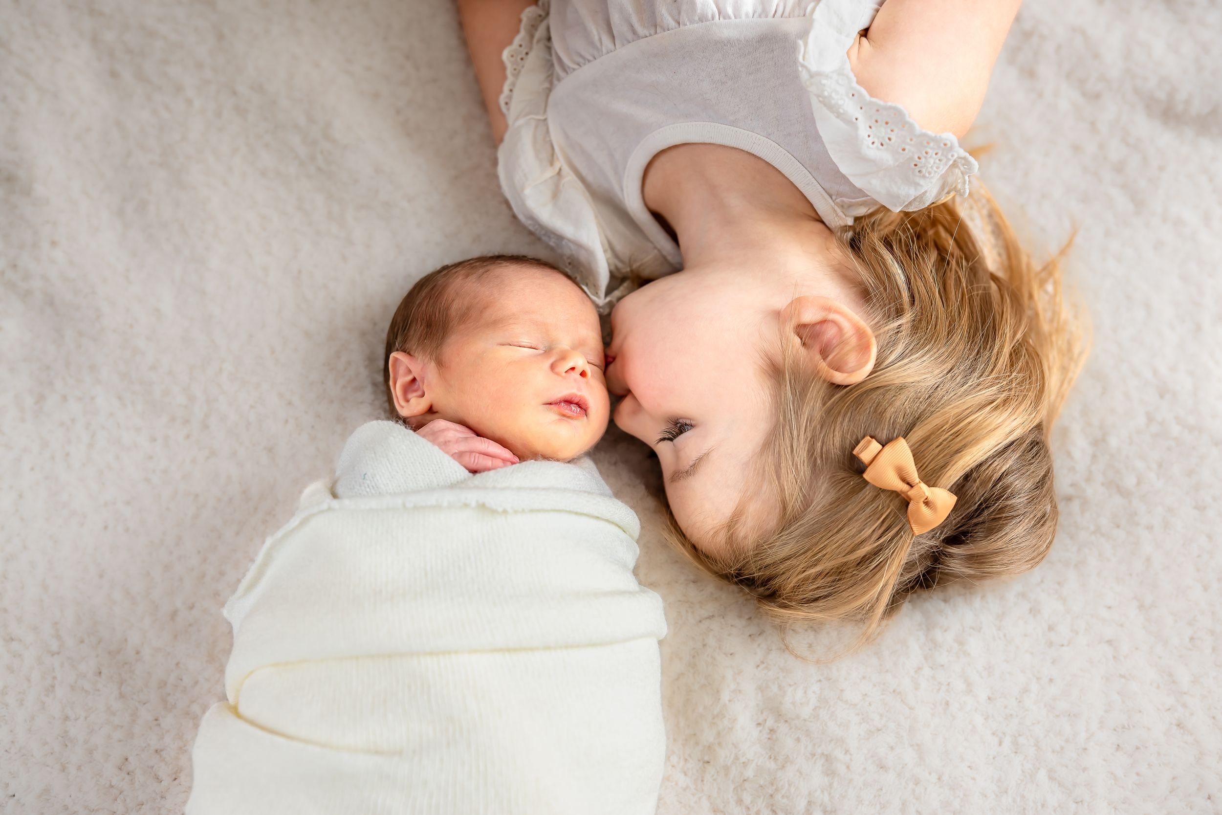 Newborn baby swaddled in white blanket lying next to smiling toddler sibling on a soft cream blanket.