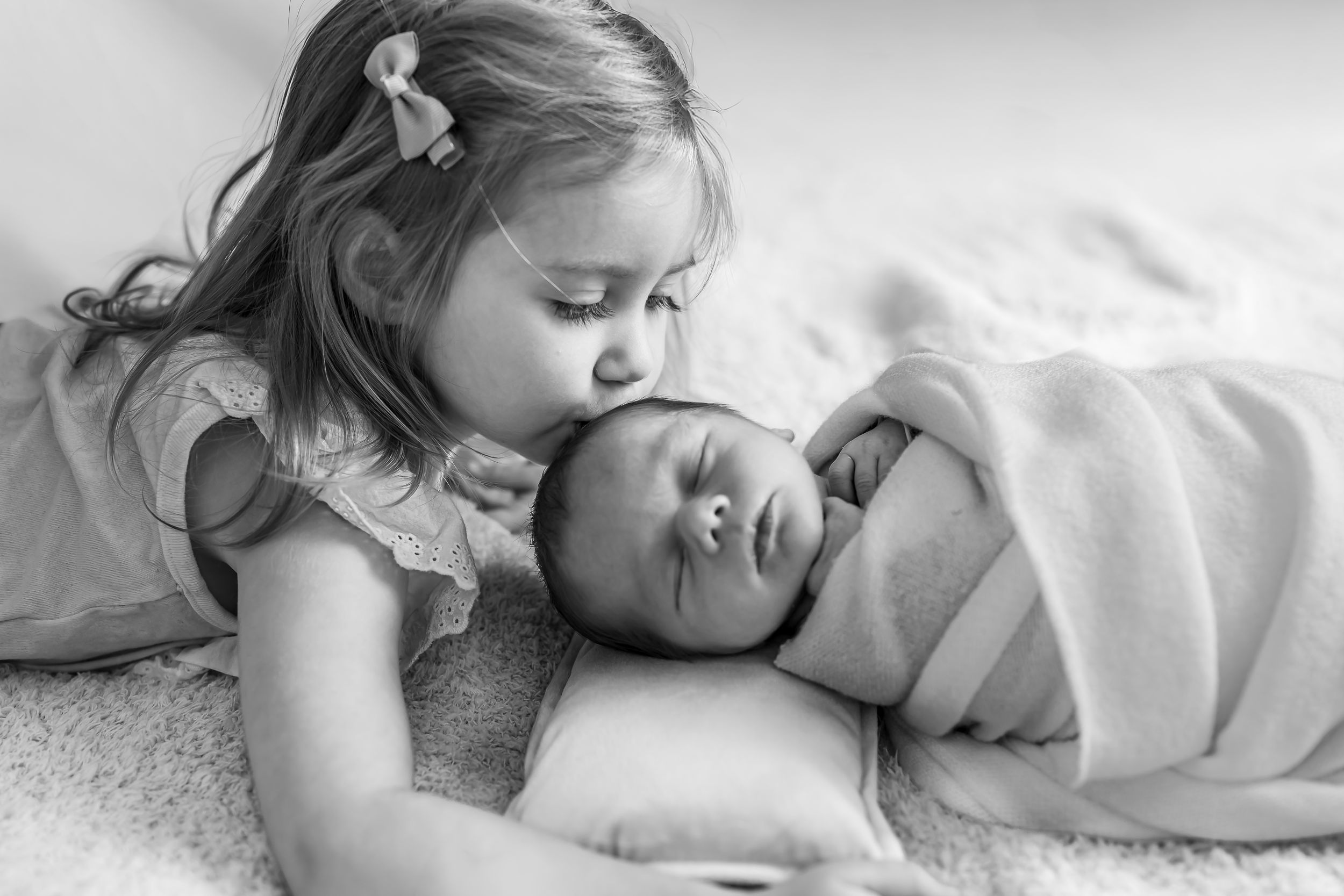 Black and white photo of a young girl kissing a sleeping newborn baby wrapped in a swaddle blanket.