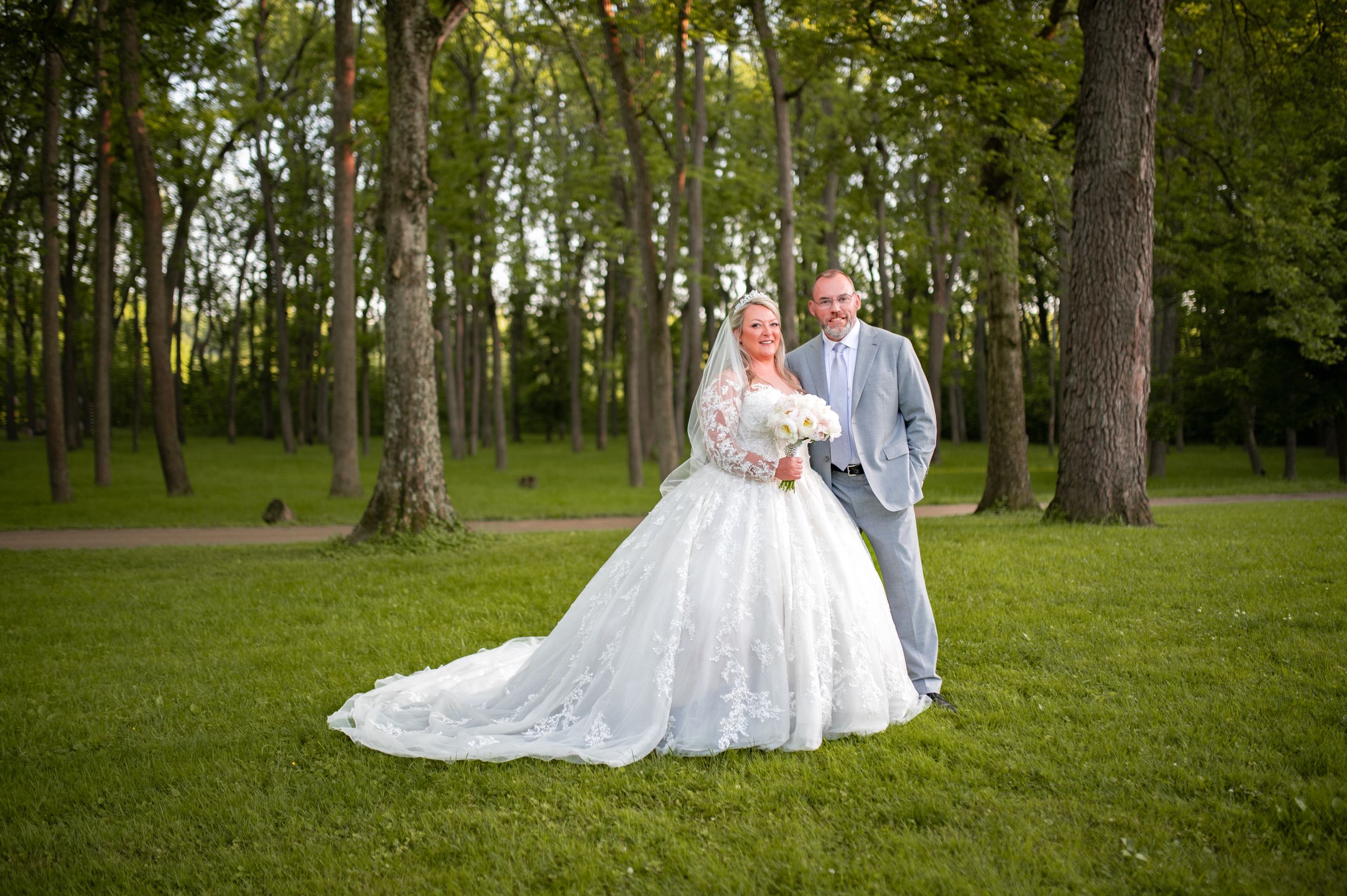 A couple in wedding attire stands together on green lawn surrounded by tall trees.