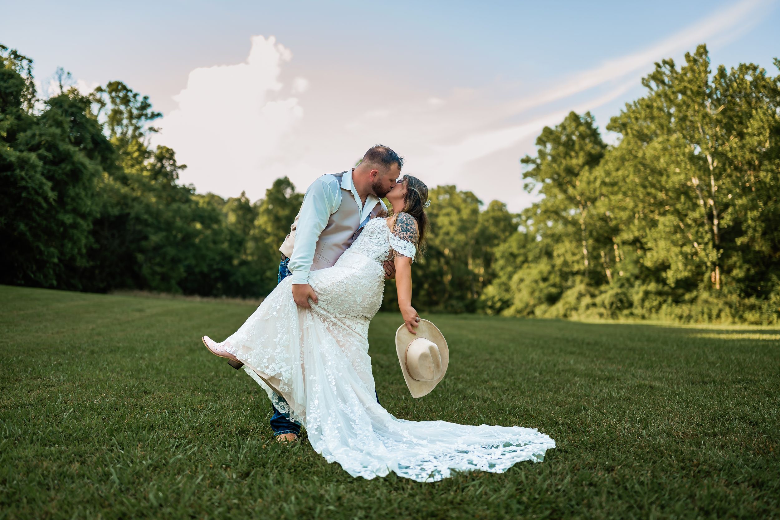 A romantic wedding couple embraces in a dip pose on a lush green lawn with trees in the background at sunset.