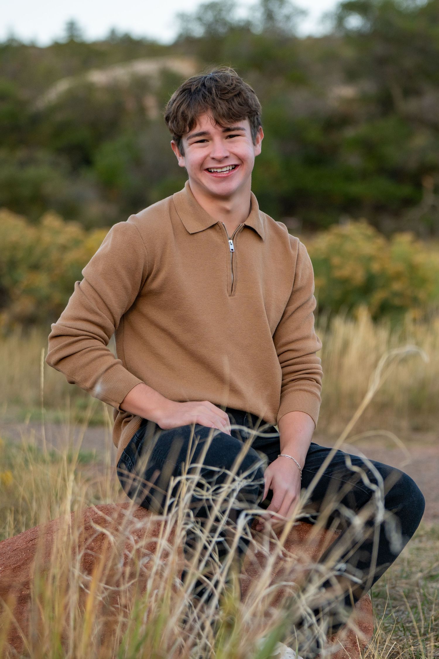 High school senior in tan sweater smiling while sitting in tall grass outdoors. Senior session