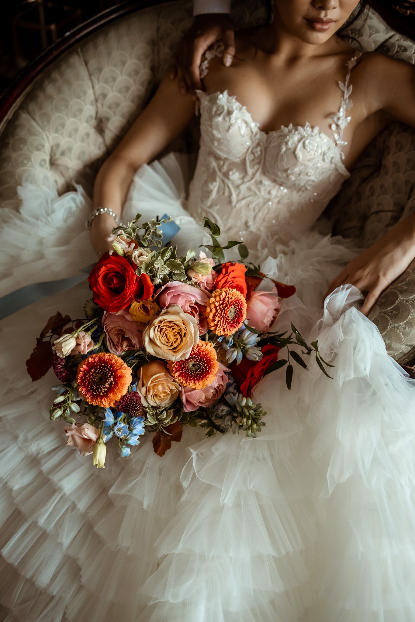 A stunning bridal bouquet with red and coral roses rests on a white tulle wedding gown with delicate lace details.