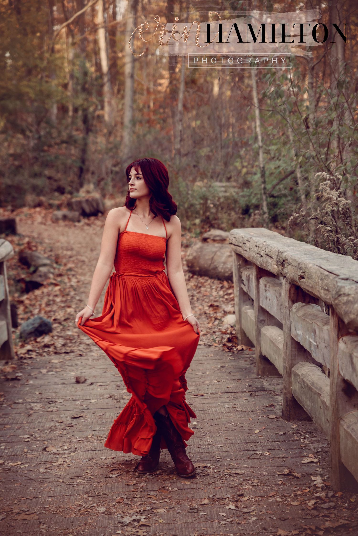 A red formal dress billows dramatically against a wooded backdrop on a wooden bridge pathway.