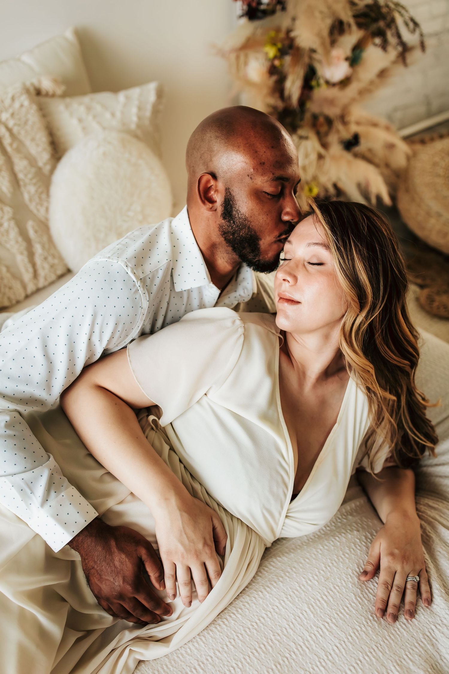 A couple shares an intimate moment while lying together on a white bed with decorative dried flowers in the background.