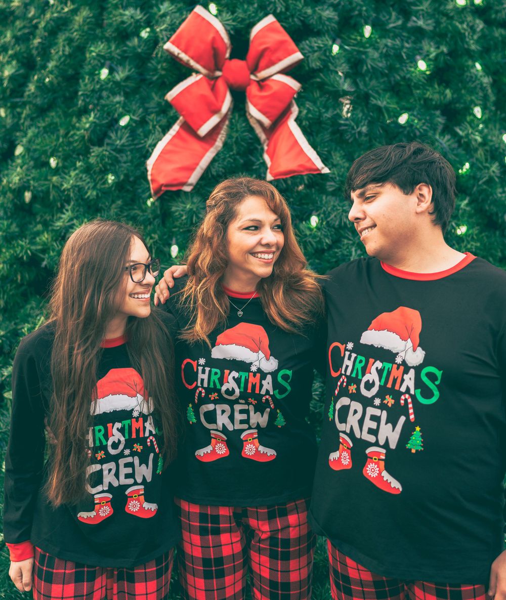 Three people wearing matching Christmas Crew holiday sweaters and plaid pajama pants pose together in front of a Christmas tree.