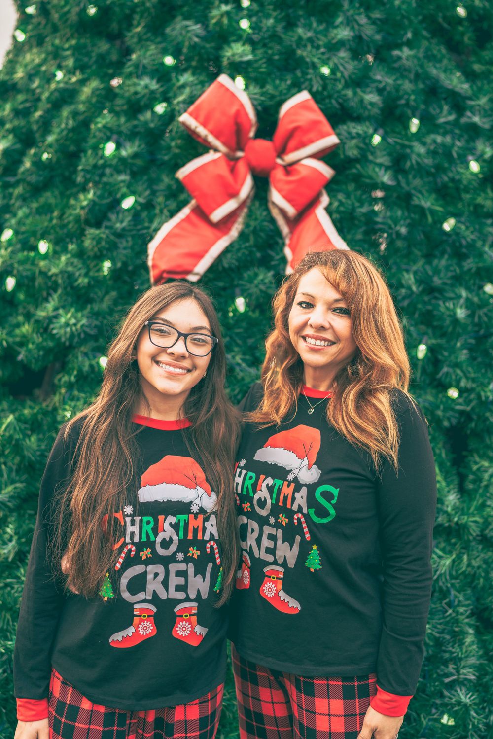 Two people wearing matching Christmas crew sweaters with plaid pants stand in front of a decorated evergreen tree.