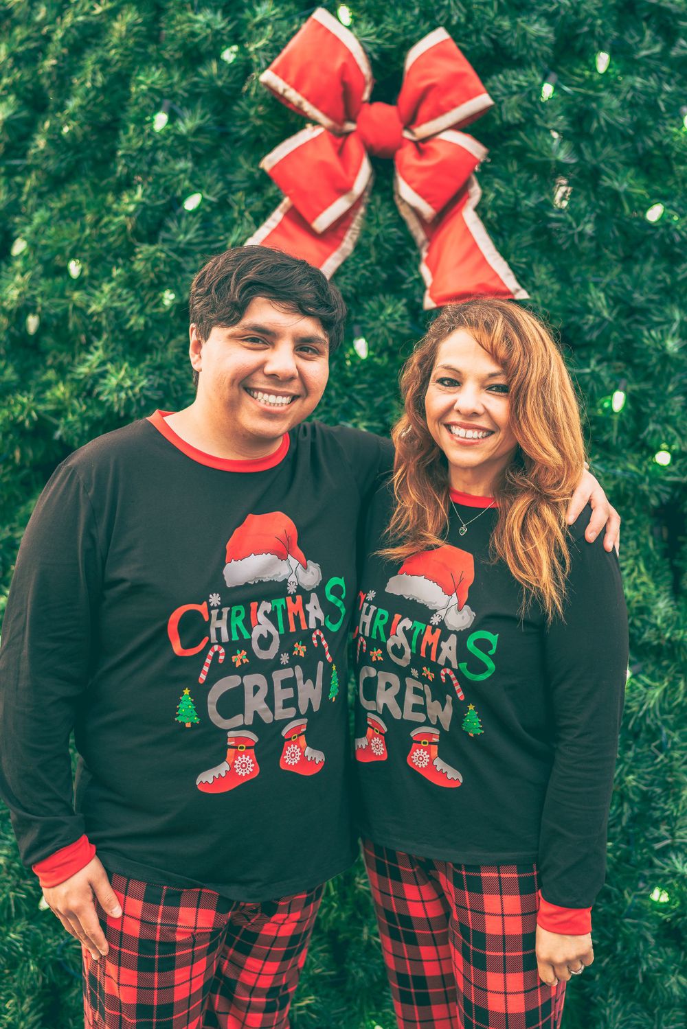 Couple wearing matching Christmas crew sweaters and plaid pants pose in front of a green wall with red bow decoration.