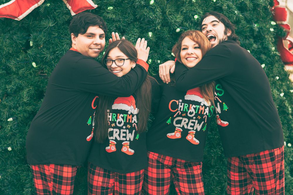 A group wearing matching buffalo plaid pajama pants and black Christmas sweaters poses together in front of evergreen trees.