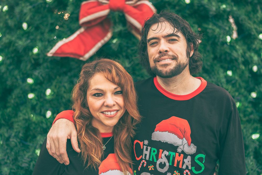 A couple wearing Christmas sweaters poses together in front of evergreen decorations with a red bow visible.