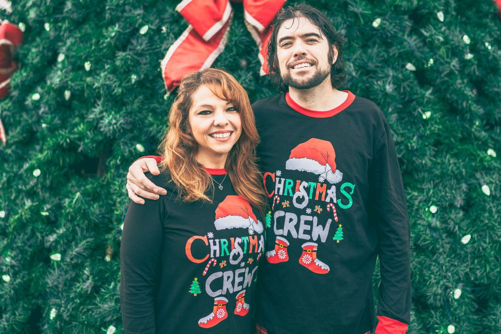 A couple wearing matching Christmas sweaters smiles together in front of evergreen foliage at wintertime.