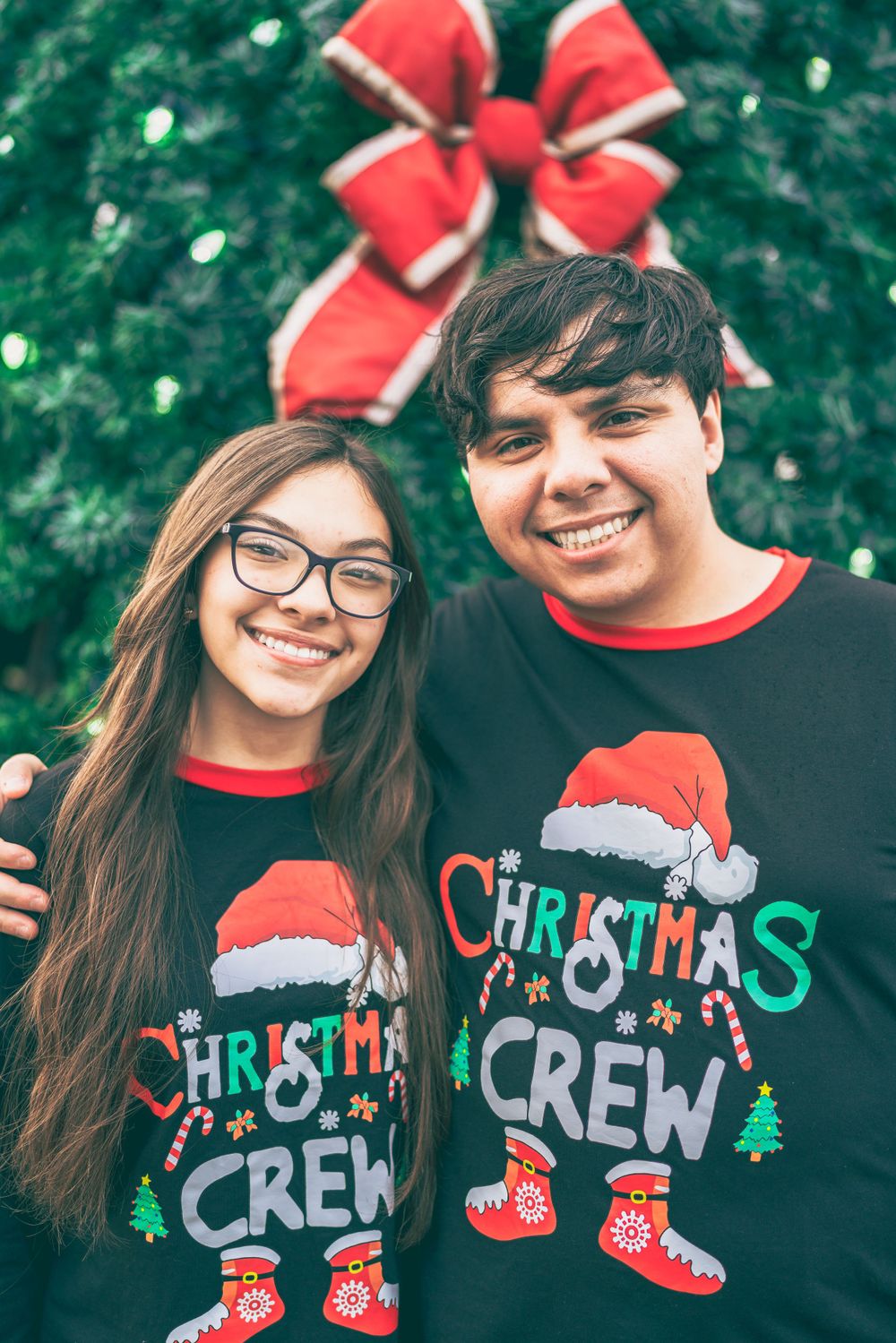 Two people wearing matching Christmas Crew sweaters pose together in front of a decorated Christmas tree with a red bow.