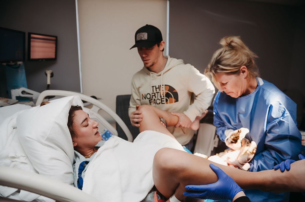Medical professional in blue scrubs assists a patient in a hospital bed during what appears to be a delivery.