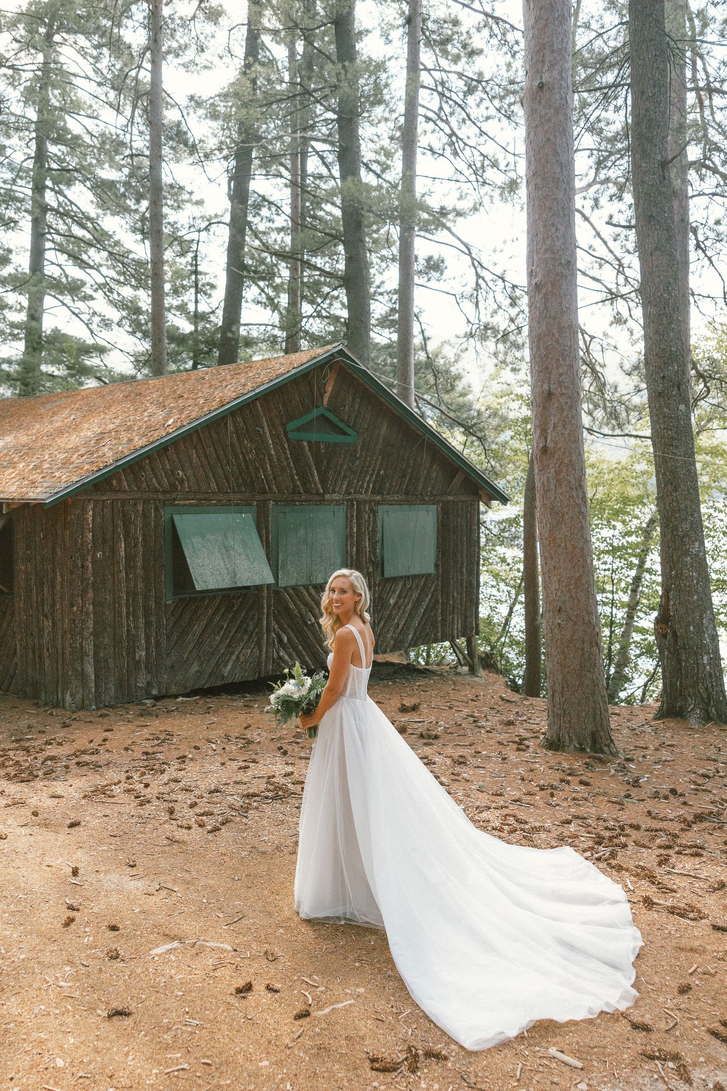 White wedding dress flowing against rustic cabin backdrop in pine forest setting.