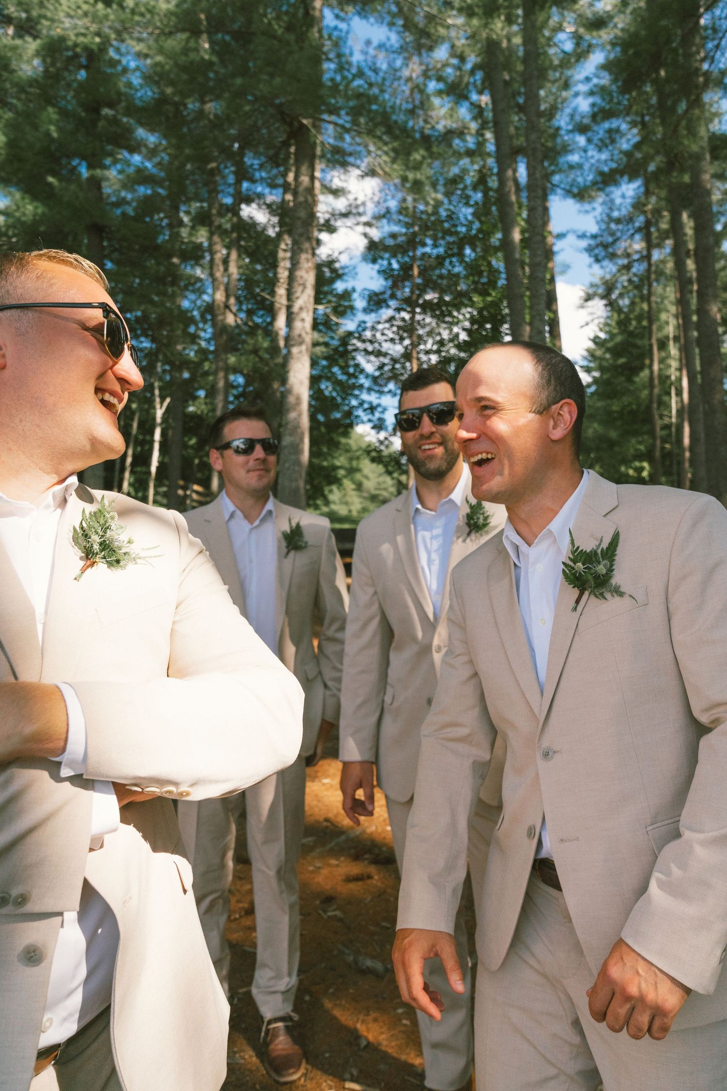 Groomsmen in beige suits and sunglasses gathered outdoors among pine trees.