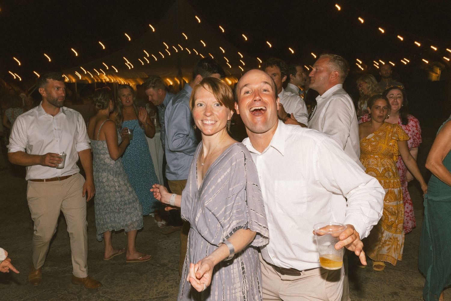 People dancing enthusiastically under warm string lights at an evening celebration.