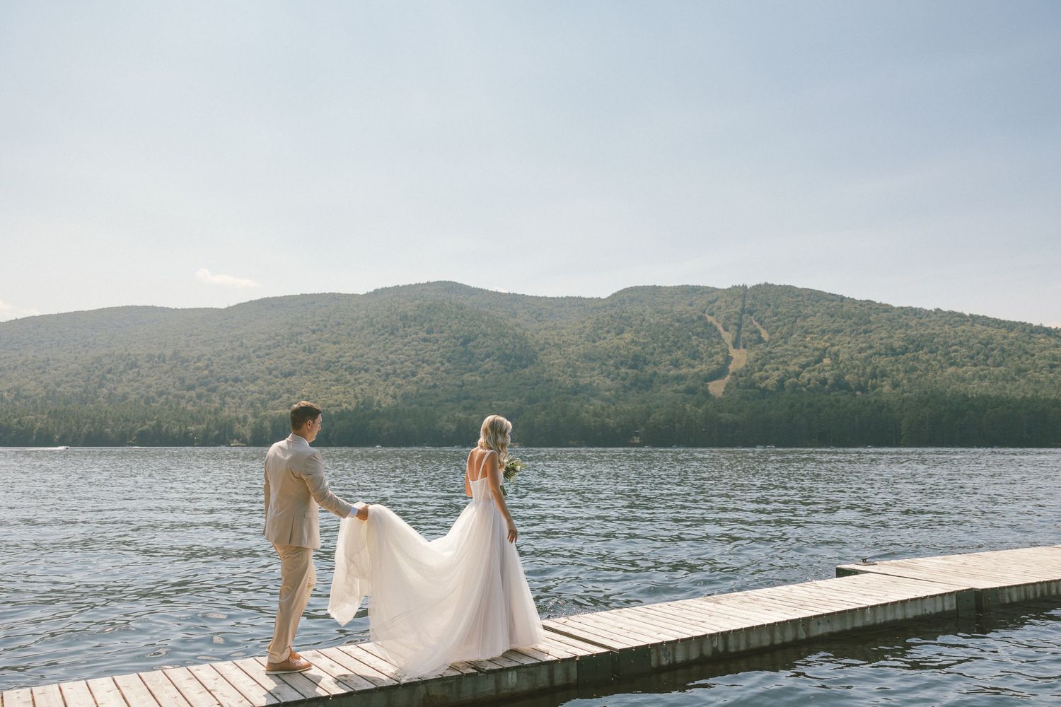 Couple standing on wooden dock overlooking mountain lake in wedding attire.