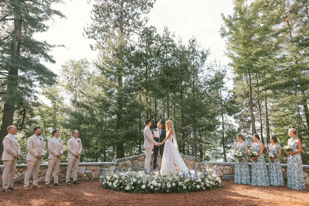 Outdoor forest wedding ceremony with floral arch and wedding party in light colored attire.