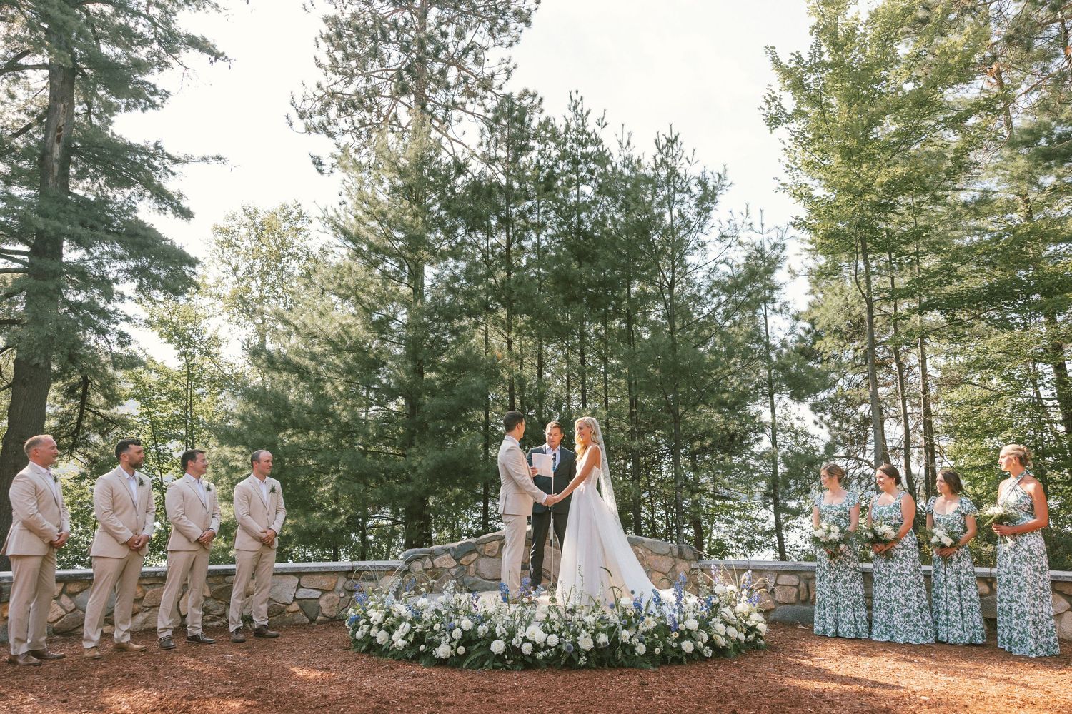 Outdoor forest wedding ceremony with floral arch and wedding party in light colored attire.