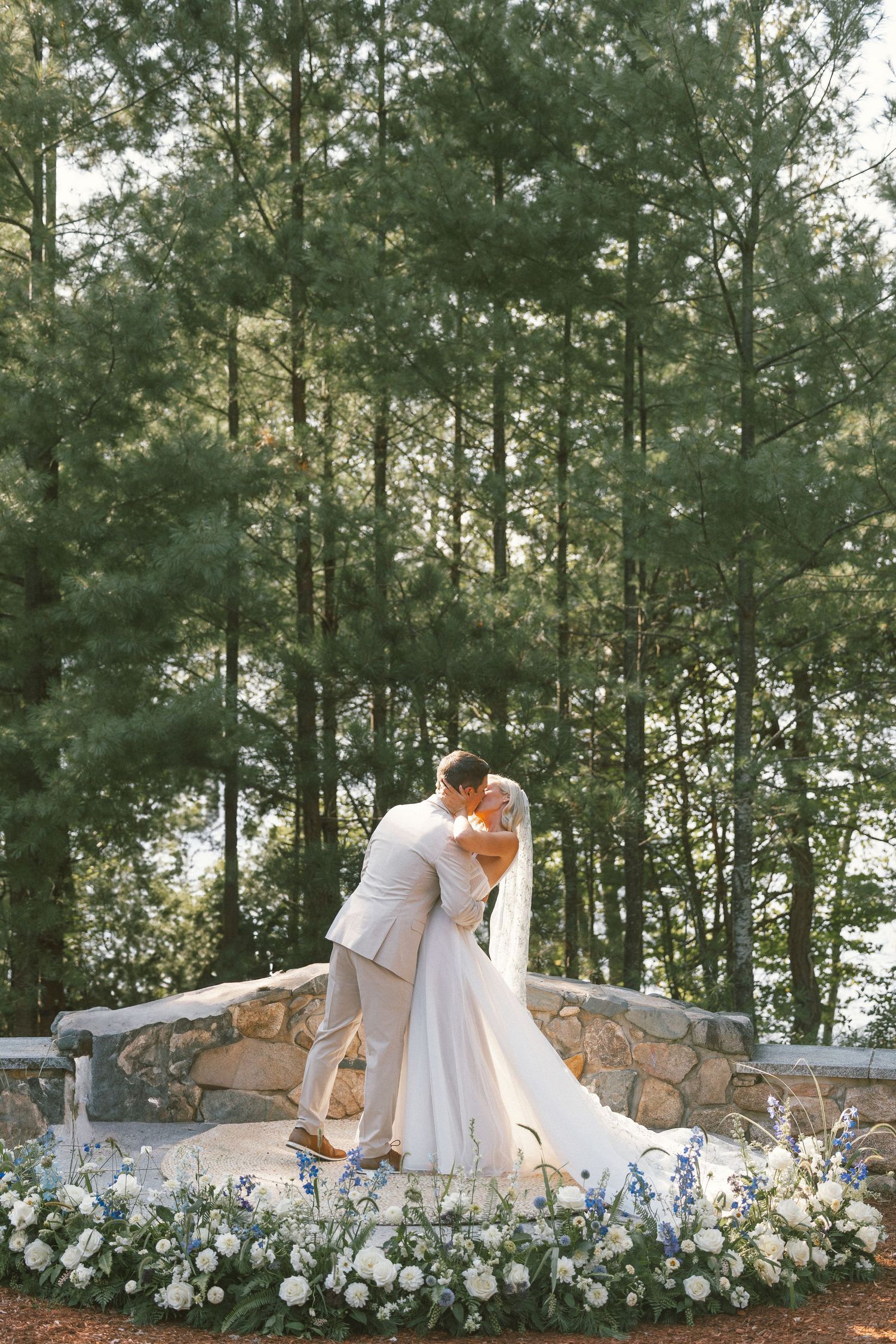 Romantic wedding couple embracing on stone platform surrounded by white floral arrangements.