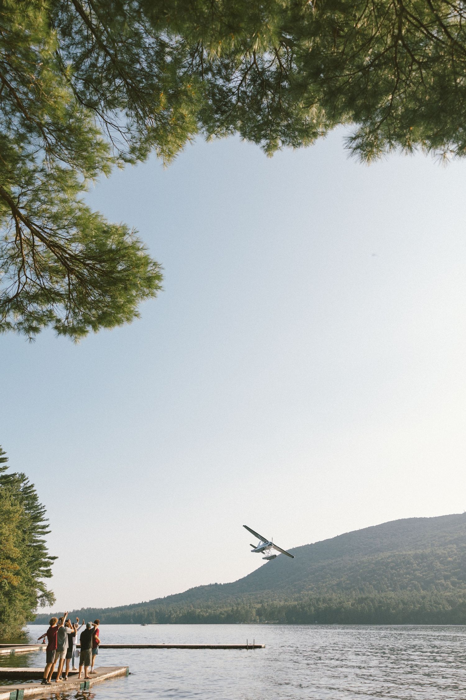 Seaplane taking off over lake with people watching from dock and mountains in background.