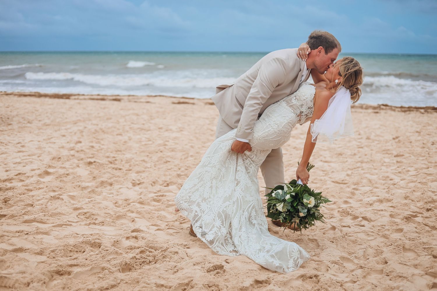 Newlyweds share a romantic kiss on a sandy beach with ocean waves in the background. Wedding photography, travel photography