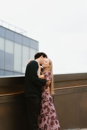 Couple embracing and kissing on a rooftop terrace, woman in a floral dress and man in dark clothes, with a modern glass building behind.