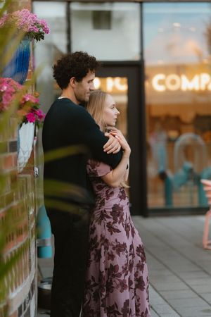 A couple embraces tenderly on a city sidewalk, the woman in a floral dress, beside pink flowers and a glowing storefront.