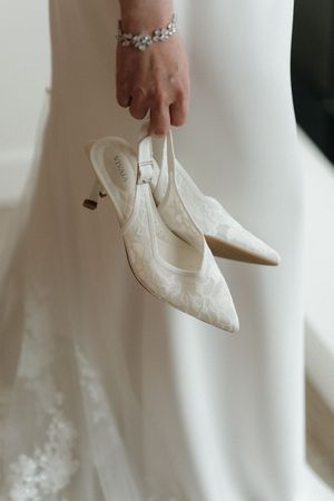 Bride in a white gown holds ivory lace kitten heel slingback wedding shoes in one hand, wearing a sparkling crystal bracelet.