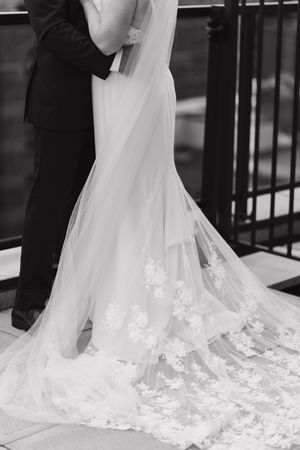 Black and white photo of a bride in a lace-appliqué wedding gown with a long sheer veil embracing a groom in a dark suit.