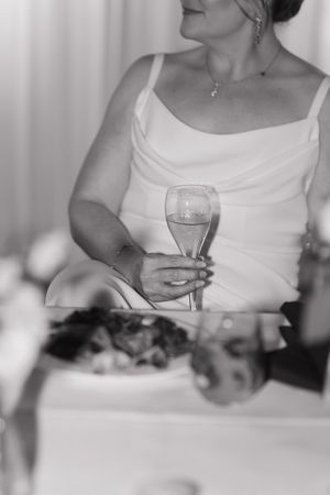 Black and white photo of a woman in a satin slip dress holding a champagne flute at a wedding table with a plate of food.