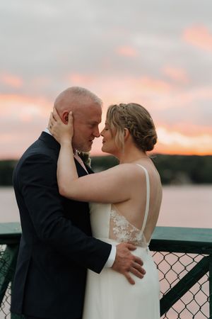 Bride and groom share an intimate moment forehead to forehead on a waterfront deck against a pink and orange sunset sky.