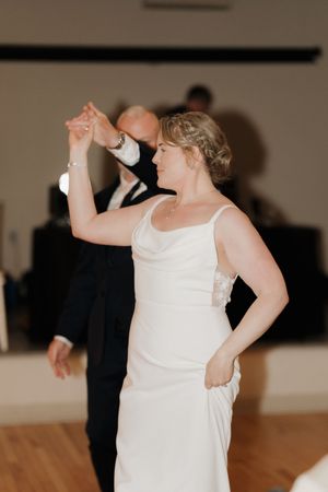 Bride in a white satin gown with an updo dances with a man in a dark suit on a wooden dance floor during a wedding reception.