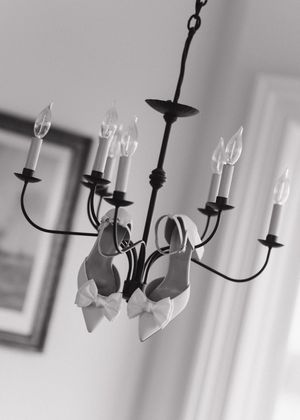 Black and white photo of bridal heels with bow details hanging from a candle-style wrought iron chandelier.
