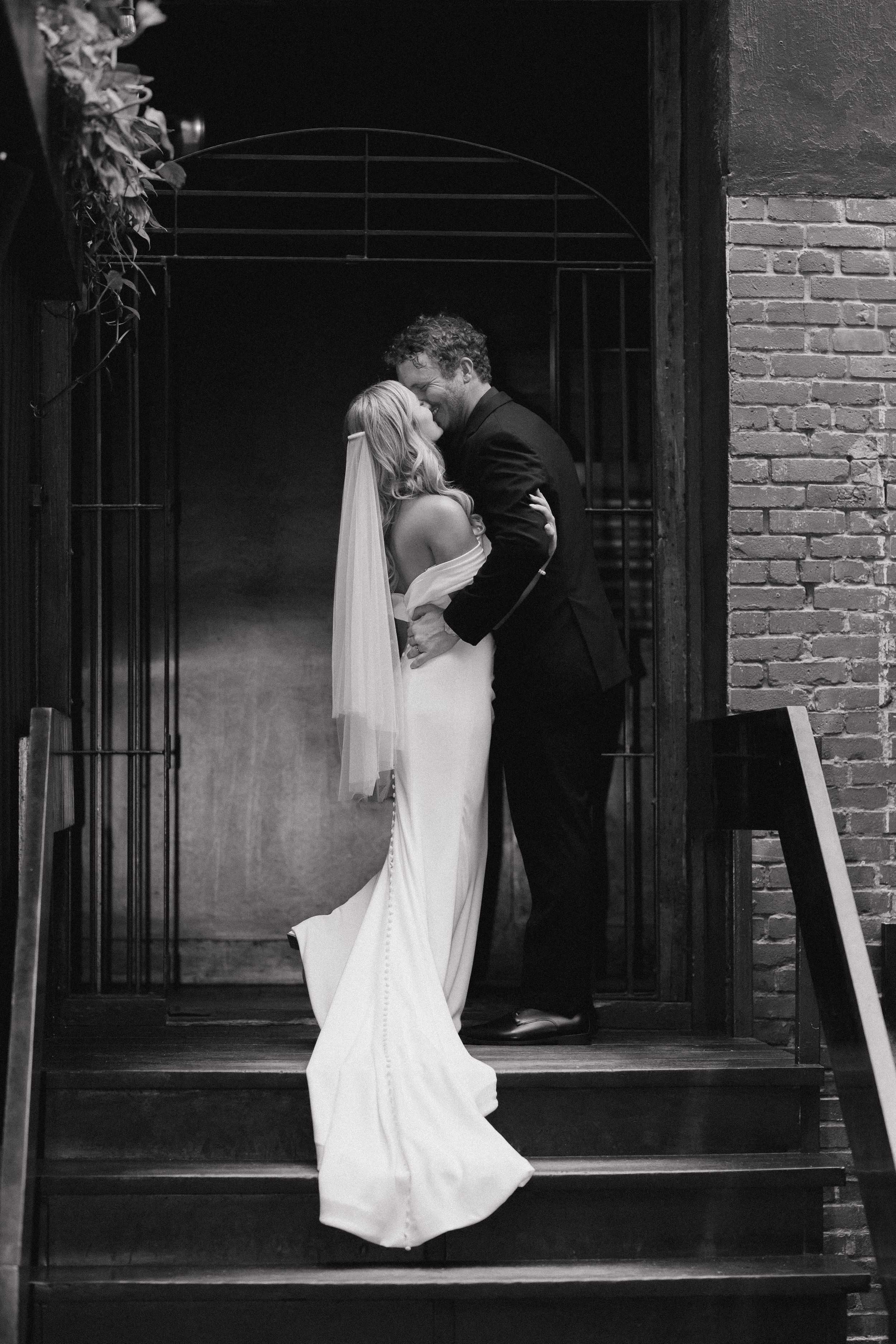 Black and white photo of a bride in a long white gown and veil kissing her groom in a dark suit on brick stairs by an iron gate.
