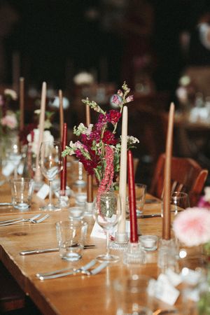 Rustic wooden banquet table styled with tall taper candles, glassware and a burgundy and pink floral centerpiece.