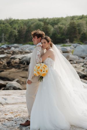 Bride in a flowing tulle gown and veil holding a yellow bouquet embraces her groom in a tan suit on a rocky coastal shore.
