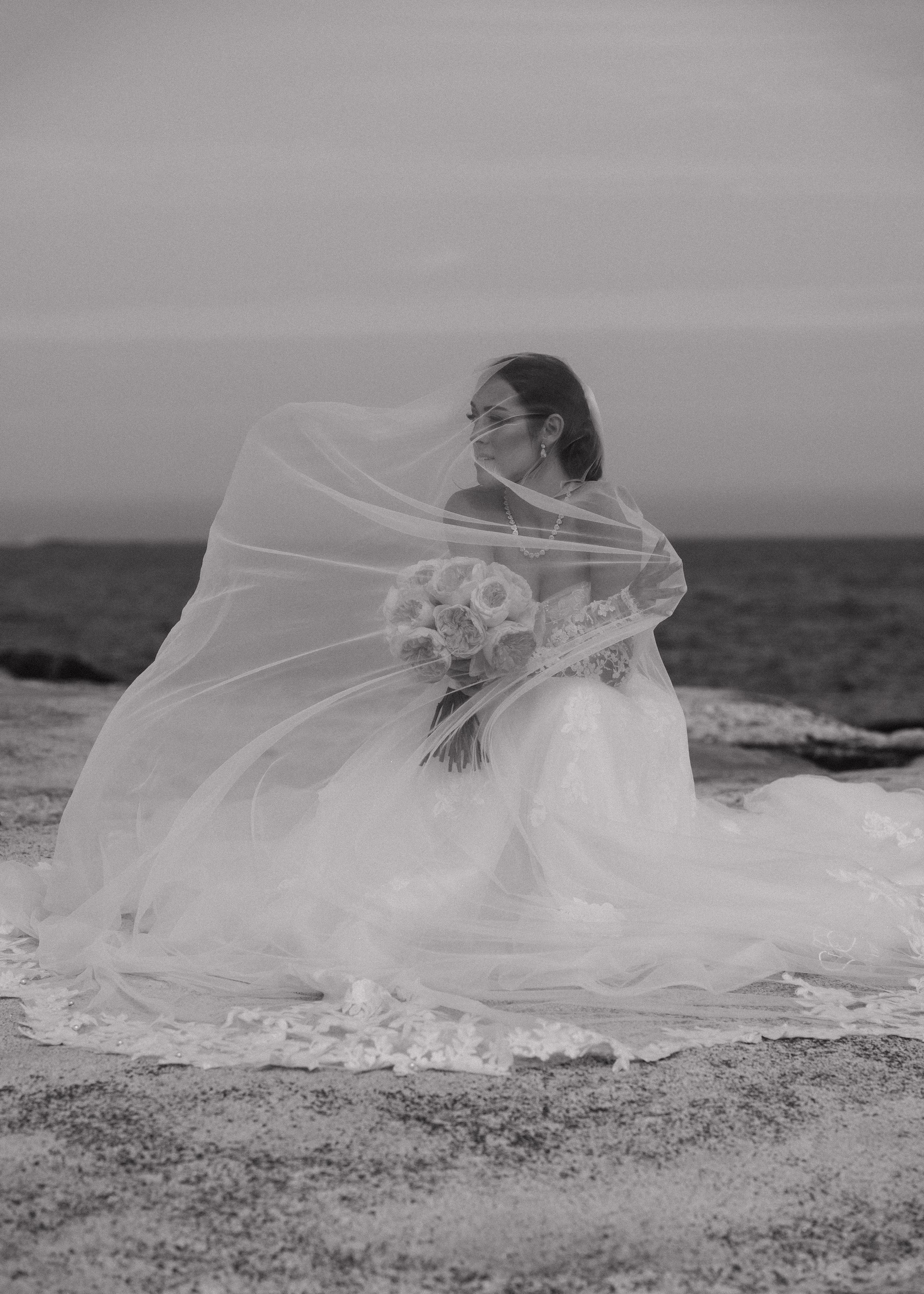 Black and white photo of a bride in a lace wedding gown holding a bouquet as the wind blows her sheer veil on a rocky seaside shore.